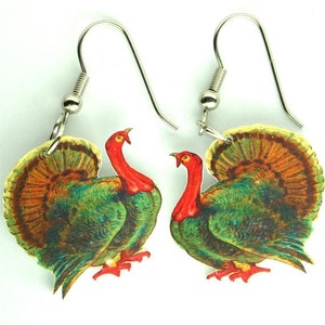 May include: A pair of dangle earrings featuring a vintage illustration of a green and brown turkey with a red head and wattle. The turkey is standing with its tail feathers fanned out.