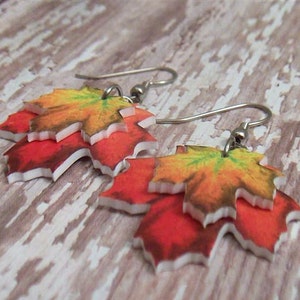 May include: A pair of earrings featuring layered maple leaves in shades of red, orange, and yellow. The leaves have a white border and are attached to silver hooks.