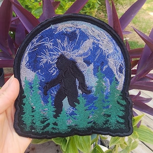May include: A black, green, and blue embroidered patch featuring a silhouette of Bigfoot standing in a forest with a full moon in the background.