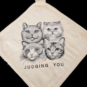 May include: Cream-colored quilted pot holder featuring four cat faces in black and white. The text "JUDGING YOU" is printed below the cat faces. The pot holder has a loop for hanging.