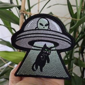 May include: An embroidered patch featuring a black cat with green eyes being abducted by a grey UFO. Inside the saucer, a pale green alien with large black eyes is visible. The design is outlined in black thread with a light green tractor beam.