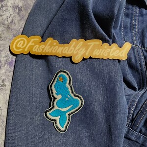 May include: A dark blue denim jacket featuring an embroidered patch of a bright blue mermaid with a small yellow star in her hair. Above the mermaid patch is a gold-colored sticker or label with the text @FashionablyTwisted in script font.