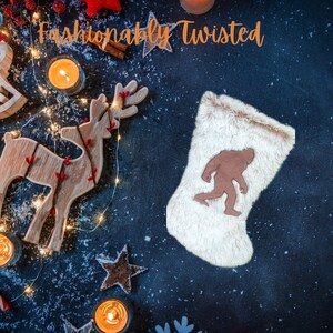 May include: A white fuzzy Christmas stocking with a brown felt Bigfoot silhouette. The stocking is surrounded by a dark blue background with a wooden reindeer, red berries, and string lights.