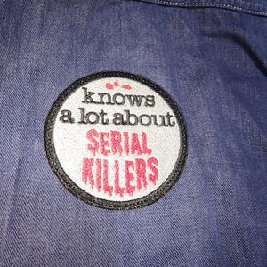 May include: A white and black circular patch with red text that reads "knows a lot about SERIAL KILLERS" on a blue denim background.