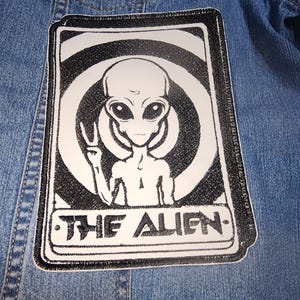 May include: Black and white embroidered patch featuring an alien with a peace sign, set against a swirling background. The text "THE ALIEN" is written below the alien.