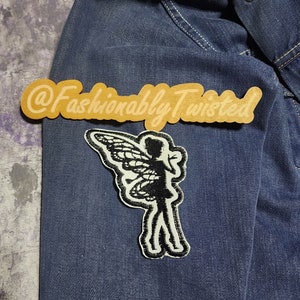 May include: A denim jacket with a black and white fairy patch. The patch features a silhouette of a fairy with detailed wings. A wooden sign with the text "@Fashionably Twisted" is also present.