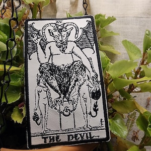 May include: Black and white embroidered patch of the Devil tarot card. The Devil is depicted as a horned, winged figure with a human-like body and goat legs. Two figures are chained to the Devil's throne.