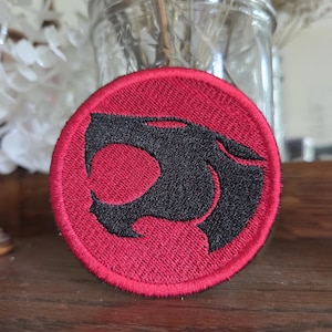 May include: A circular embroidered patch featuring a black stylized cat head silhouette centered on a vibrant red background. The patch has a thick, stitched red border that defines its round shape, with dense thread work throughout the design.