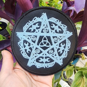 May include: A black leather patch with a white embroidered pentagram with a triple moon symbol in the center. The pentagram is surrounded by a decorative border.