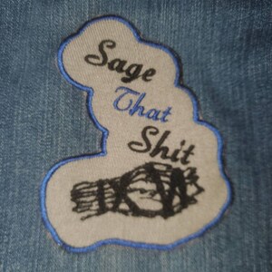 May include: Sage That Shit embroidered patch with a hand holding a bundle of sage. The patch is white with blue stitching and is on a blue denim background.
