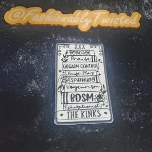 May include: A rectangular, clear acrylic charm with black text listing BDSM-related terms, including "Bondage," "Praise," and "The Kinks." The charm has floral and star accents and the text "@Fashionably Twisted" in an orange, cursive font.