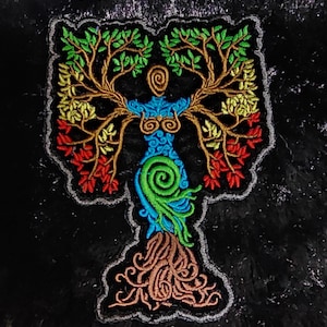 May include: Embroidered patch featuring a stylized tree of life design. The patch depicts a figure with a blue torso, green spiral, and brown roots, with branches of green, yellow, and red leaves. The patch is outlined in gray on a black background.