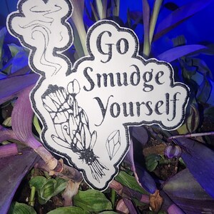 May include: A white sticker with black border and text that reads "Go Smudge Yourself." The sticker features a drawing of a bundle of herbs, a crystal, and smoke. The sticker is set against a background of green and purple plants.