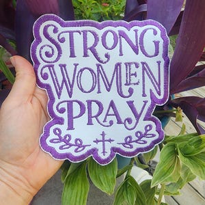 May include: Embroidered patch with the words "STRONG WOMEN PRAY" in purple thread on a white background. The patch has a decorative purple border and a cross design. The patch is held in front of a plant with purple and green leaves.