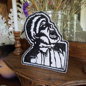 May include: A black and white embroidered patch featuring a stylized graphic of a person with their head tilted back and mouth open in an expressive pose. The design uses thick black thread for the outlines and shading against a white fabric base.