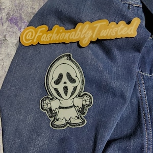 May include: A blue denim jacket with a white and black Ghostface character patch and a gold sticker that says "@Fashionably Twisted" on the left sleeve.