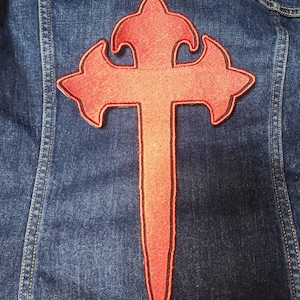 May include: A red embroidered cross patch with a pointed bottom, sewn onto a blue denim jacket.