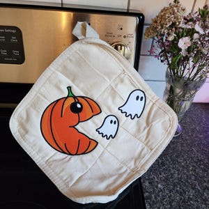 May include: Cream-colored quilted pot holder with a Halloween-themed design. An orange pumpkin with a black eye and mouth is chasing two white ghosts. The pot holder has a loop for hanging.