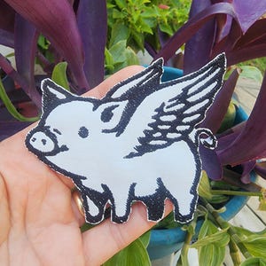 May include: White embroidered iron-on patch of a cartoon pig with wings. The pig has a black outline and black eyes. The wings are detailed with black stitching.
