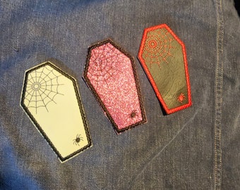 May include: Three coffin-shaped patches with Halloween themes. One is cream-colored with a spiderweb design, another is pink with glitter, and the third is black with a red spiderweb design. Each patch has a spider and black trim.