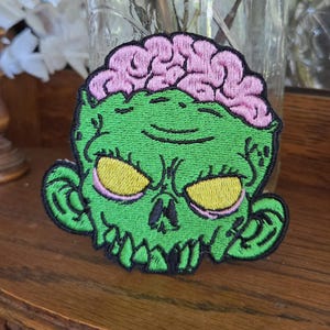 May include: An embroidered patch featuring a green zombie head with a prominent pink brain exposed on top. The zombie has yellow eyes, a stitched nose, and sharp teeth, all outlined in black thread against a vibrant green background.