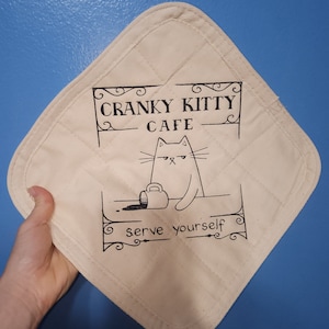 May include: A white cotton pot holder with a black and white illustration of a grumpy cat with a coffee cup that has spilled on the counter. The text reads "Cranky Kitty Cafe" and "serve yourself".
