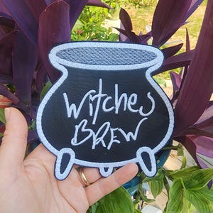 May include: Black cauldron-shaped patch with white stitching. The patch says "Witches Brew" in white cursive lettering.