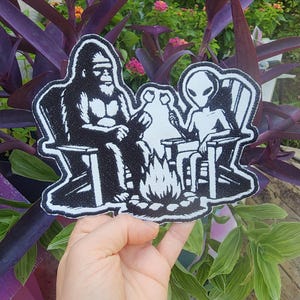 May include: Black and white embroidered patch featuring a Bigfoot and an alien sitting around a campfire. The patch is on a hand.