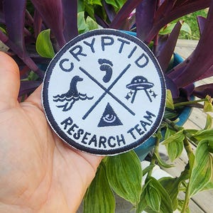 May include: White round patch with black embroidered text that reads "Cryptid Research Team". The patch features a footprint, a UFO, a sea monster, and an all-seeing eye.