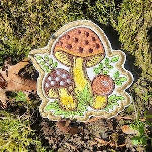 May include: A brown, white, and yellow embroidered patch of three mushrooms with green leaves. The patch is on a brown and green mossy background.