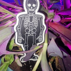 May include: A white embroidered skeleton figure on a black background. The skeleton is standing in a pot of green and purple plants.