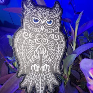 May include: A gray embroidered owl patch with white thread detailing. The owl has blue eyes and a detailed design on its feathers.