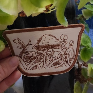 May include: A brown embroidered patch with a white background featuring a group of mushrooms with a brown outline. The patch is shaped like a rounded rectangle with a brown border.