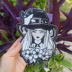 May include: Black and white embroidered patch of a witch with a hat, long hair, and flowers. The patch is on a black background.