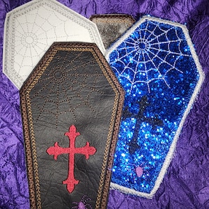 May include: Three embroidered patches in the shape of coffins. The first is white with a spiderweb design, the second is brown with a cross and spider, and the third is blue with glitter and a spiderweb design.