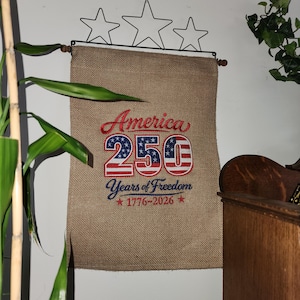 May include: A burlap banner with the text "America 250 Years of Freedom 1776-2026" in red, white, and blue. The banner is held by a decorative metal star display. The banner is hanging on a white wall.