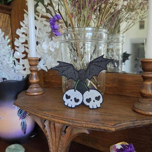 May include: An embroidered patch featuring two light gray skulls with heart-shaped eye sockets, connected by green stems to a pair of black bat wings. The design is rendered in a flat, stitched style with dark outlines, creating a cohesive graphic.