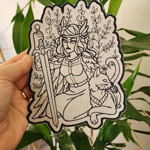 May include: A black and white embroidered patch featuring a warrior woman with a sword, a cat, and floral elements. The patch has a black border and is set against a background of green foliage.