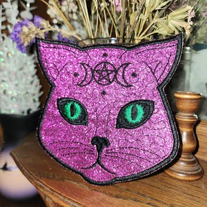 May include: A glittery purple cat-shaped patch with green eyes and a black outline. The patch features a crescent moon and star design above the eyes. The patch is on a wooden surface.