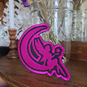 May include: A hot pink embroidered patch featuring a silhouette of a fairy sitting on a crescent moon. The design is outlined in black thread, creating a sharp contrast against the vibrant pink color of the fairy and the moon shape.