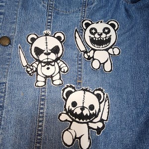 May include: Three white and black embroidered patches of teddy bears holding knives are displayed on a blue denim jacket. Each bear has a different facial expression, with one crying, one smiling, and one with a menacing grin.