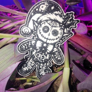 May include: Black and white embroidered patch of a skeleton elf wearing a Santa hat. The elf is holding a candy cane and has a stitched-up smile.