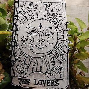 May include: A rectangular white patch featuring black embroidered line art of a sun and moon face design. The text THE LOVERS is embroidered in black at the bottom. The patch is suspended by a black metal chain and has a stitched border.