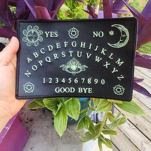 May include: Black felt Ouija board with green lettering. The board features a traditional layout with letters, numbers, and the words "YES", "NO", and "GOOD BYE".