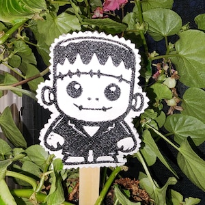 May include: Black and white cartoon Frankenstein character with stitches on his neck, wearing a black jacket, on a wooden stick. The character is standing in front of a green leafy plant.