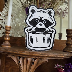 May include: A black and white embroidered patch featuring a cartoon raccoon peeking out from inside a trash can. The design uses bold black outlines and solid black fill for the raccoon's mask and paws, contrasted against a white background.