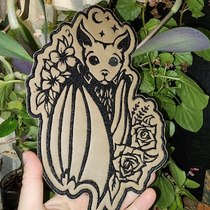 May include: A tan and black bat-shaped patch with floral accents. The patch features a bat with a crescent moon and stars above its head, surrounded by flowers and roses. The patch is held in a hand.