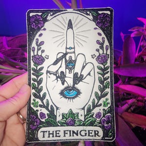 May include: Embroidered patch featuring a hand with the middle finger extended. The design includes an eye on the palm and is framed by purple flowers and green leaves. The text "THE FINGER" is at the bottom.