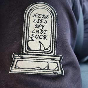 May include: Embroidered patch in the shape of a tombstone with the words "HERE LIES MY LAST FUCK" in black script. The patch is white with a black border and is sewn onto a dark gray fabric.