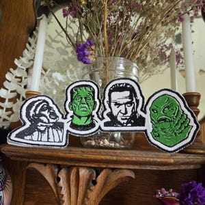 May include: A horizontal embroidered patch featuring four distinct horror character portraits. The design includes a black and white werewolf, a green and black Frankenstein's monster, a black and white vampire, and a green and black gill-man creature.